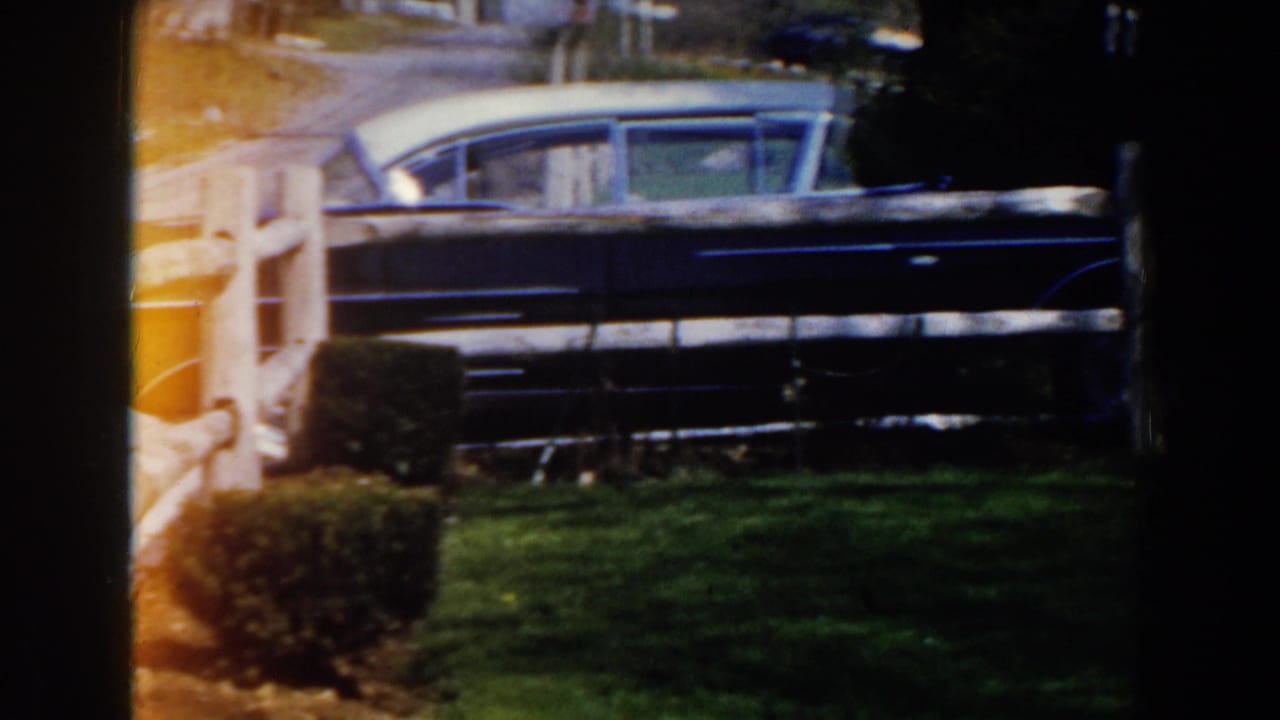 Blue sedan parked behind a white wooden fence beside a green lawn.