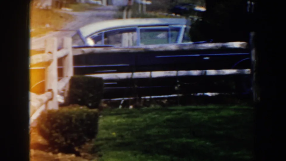 1959 Michigan home-movie frame showing a blue sedan beyond a white wooden fence, with lawn, shrubs, and trees in sunlight.