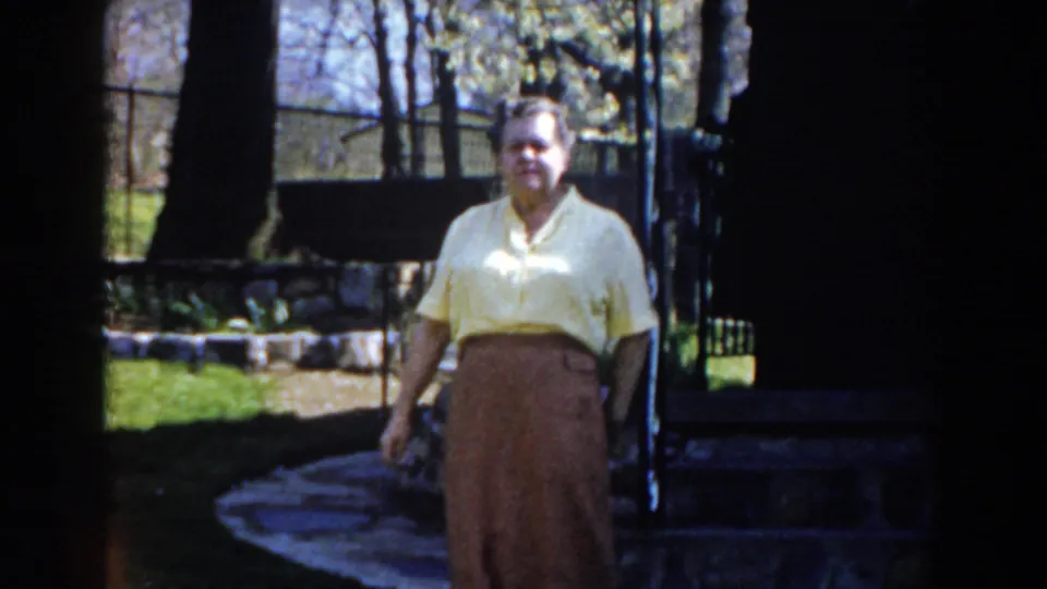 1959 Michigan home-movie frame showing an adult woman walking through a yard near a path, trees, lawn, and a fence or gate.