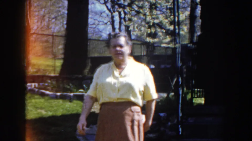 1959 Michigan frame showing an adult woman in a yellow blouse and brown skirt walking near trees, lawn, and a dark gate or fence.