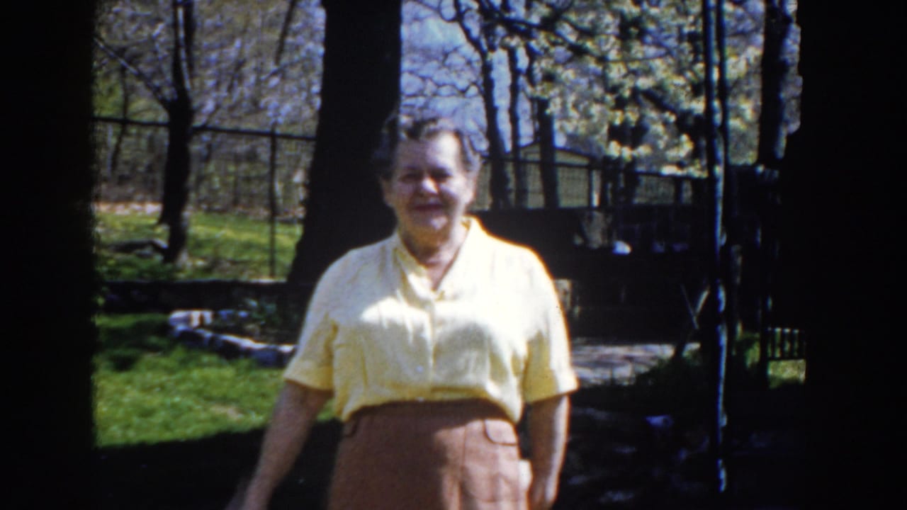 In 1959 Michigan home-movie footage, a mature woman in a yellow blouse and brown skirt faces the camera in a sun-dappled backyard.