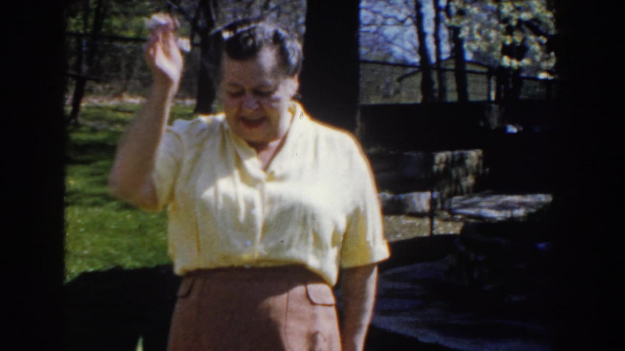 Woman in a yellow blouse raises one hand while standing outside near trees and yard structures.