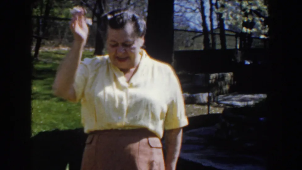 The mature woman raises her hand toward the camera in the Michigan backyard, with dark tree trunks and lawn in the background.