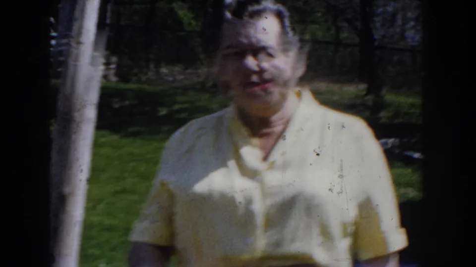 A closer 8mm frame shows the woman approaching the camera, her face softened by focus and film wear, with lawn and fencing behind her.
