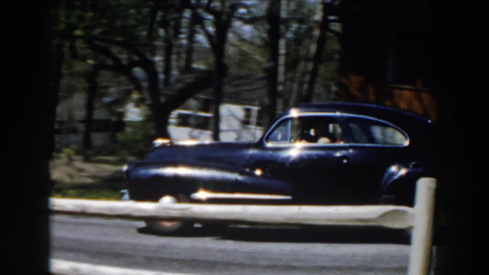 A dark 1950s-style car passes on a road in Michigan, framed by trees, fencing, and a residential-looking outdoor area.