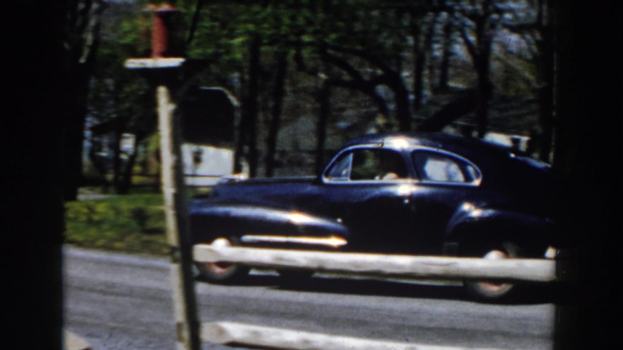Dark blue vintage sedan passing a roadside mailbox on a tree-lined street.