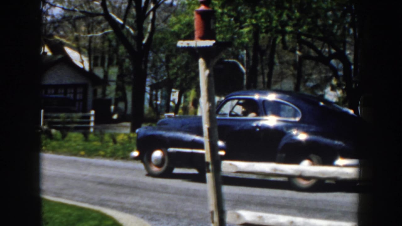 The blue sedan continues past a red mailbox on a quiet Michigan suburban street, with houses and trees in the background.