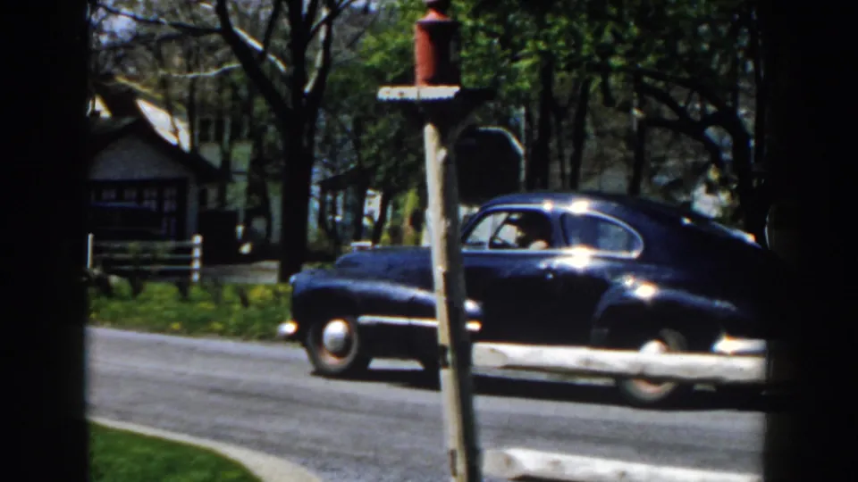 The blue sedan continues past a red mailbox on a quiet Michigan suburban street, with houses and trees in the background.