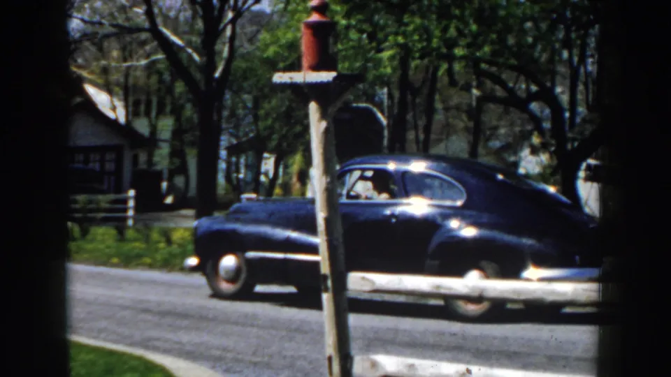 A 1959 home-movie frame shows the blue sedan nearer the camera, partly obscured by the mailbox post on a Michigan street.