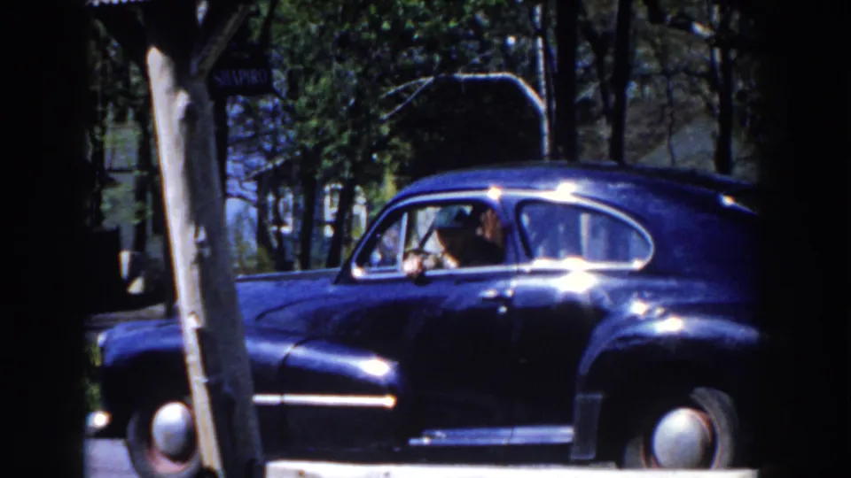 The sedan fills more of the frame, with a visible occupant through the side window and bright sunlight reflecting from the paint.