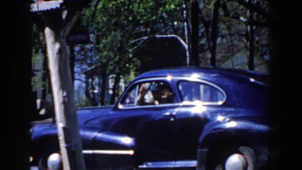 1959 Michigan home-movie frame showing a blue sedan near trees, with people visible through the side window.