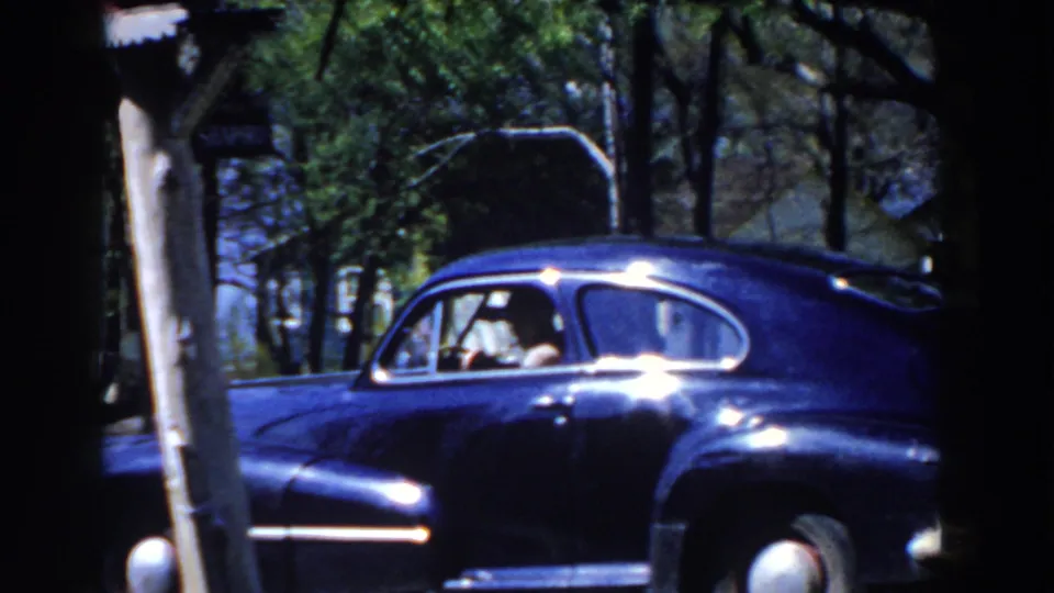 1959 Michigan home-movie frame of a blue sedan partly blocked by a foreground post, with occupants inside.