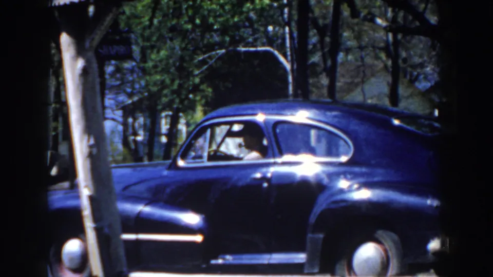 1959 Michigan frame showing a blue vintage sedan by trees as an occupant leans toward the side window.