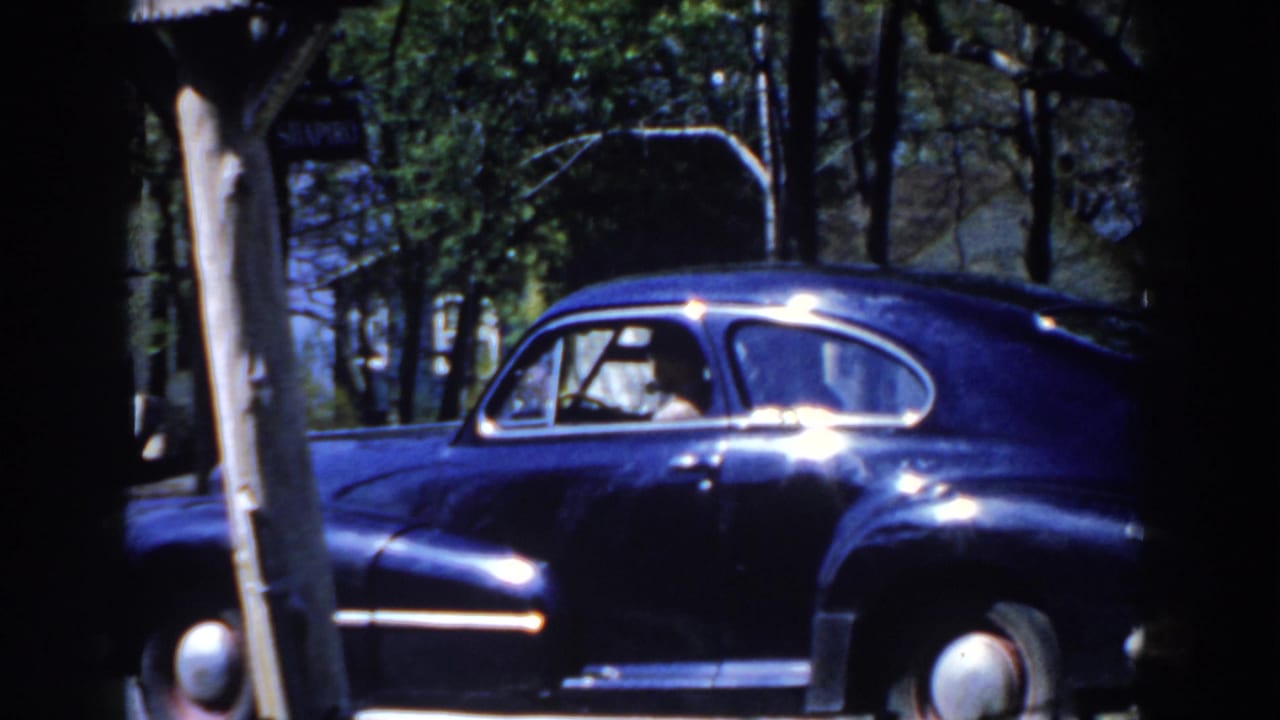 Dark blue classic sedan passing behind trees on a residential street.