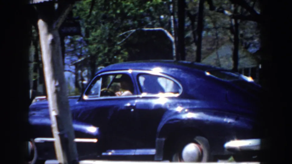 A hat-wearing driver appears through the side window as the dark blue classic car passes a pole, street sign, and leafy residential background.