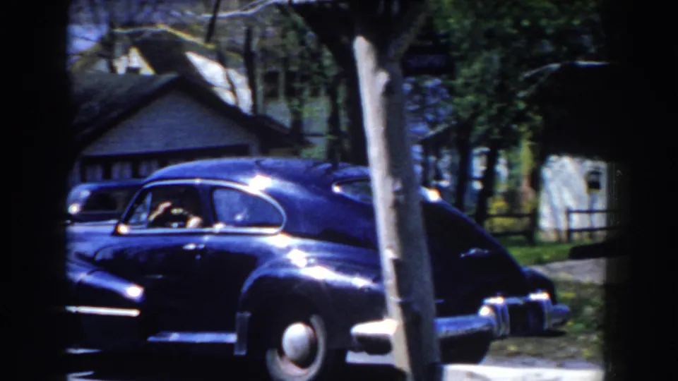 The sedan moves farther along the residential street, with its rear body, chrome bumper, a pale pole, trees, and nearby houses visible.
