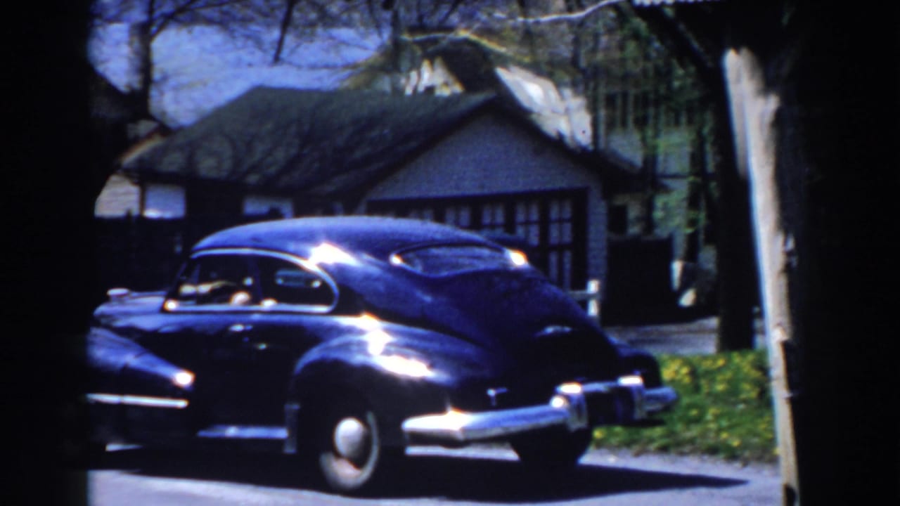 Black vintage sedan passing a white house and trees on a sunny residential street.