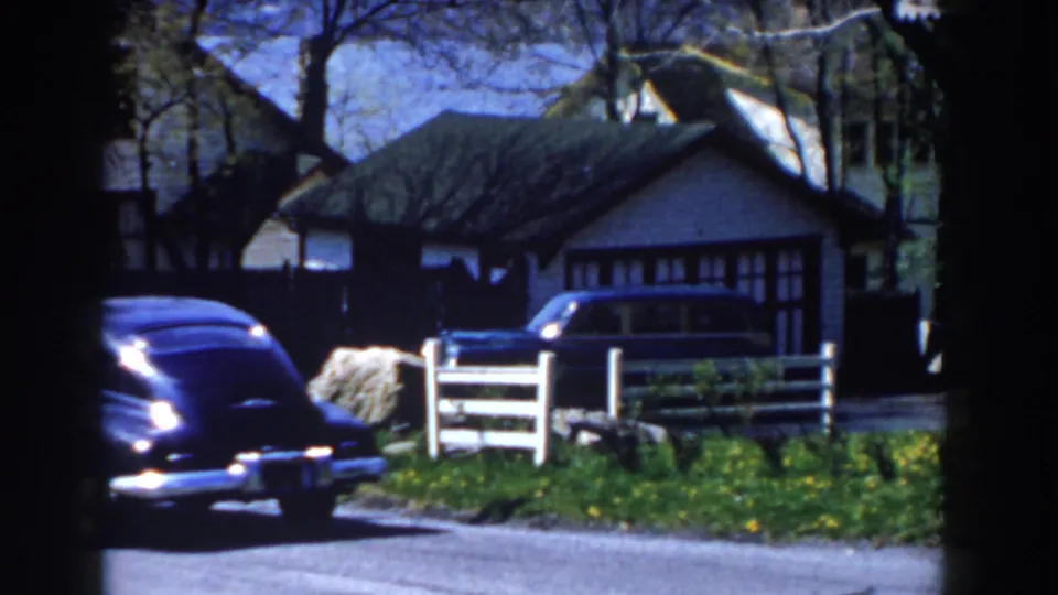 The black sedan continues along the driveway area near a white fence, residential lawn, and house in Michigan 1959 footage.