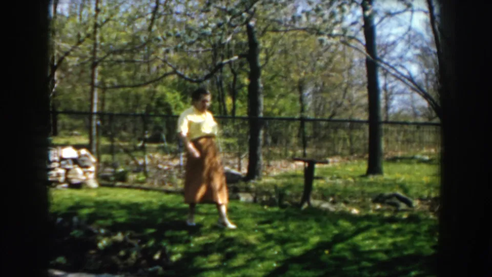 The woman remains in the grassy yard, framed by trees, a fence, stone edging, and spring foliage in Michigan 1959 home-movie footage.