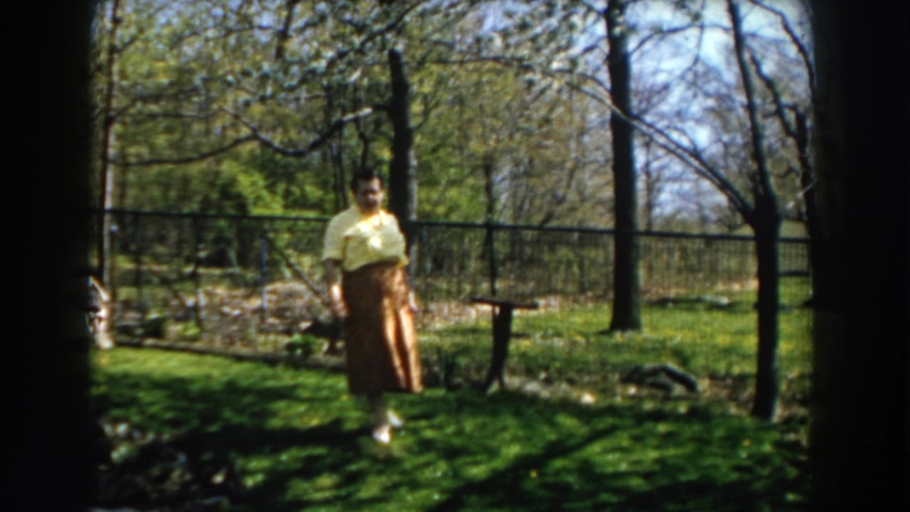 Woman standing on a green backyard lawn near garden beds and trees.