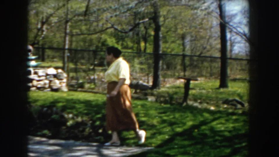 The woman moves in profile past the patio edge, with lawn, trees, fence, and garden plantings visible in the yard.