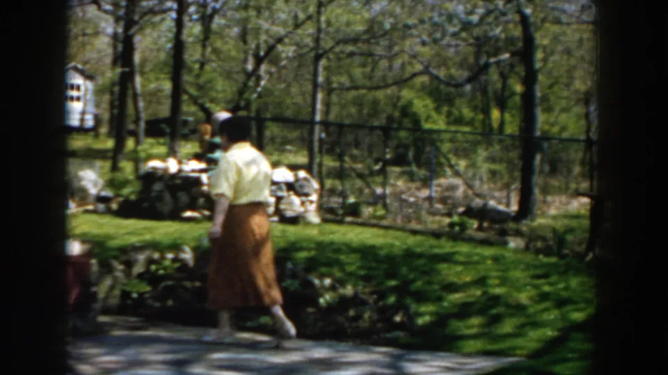 The woman walks away through the backyard garden, framed by trees, a fence, stone edging, and springlike greenery.