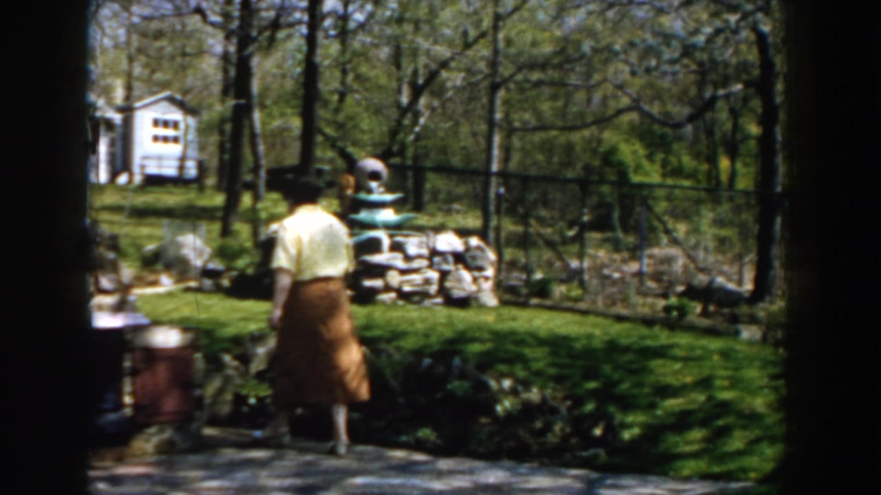 Woman walking across a sunny backyard toward a garden fountain.