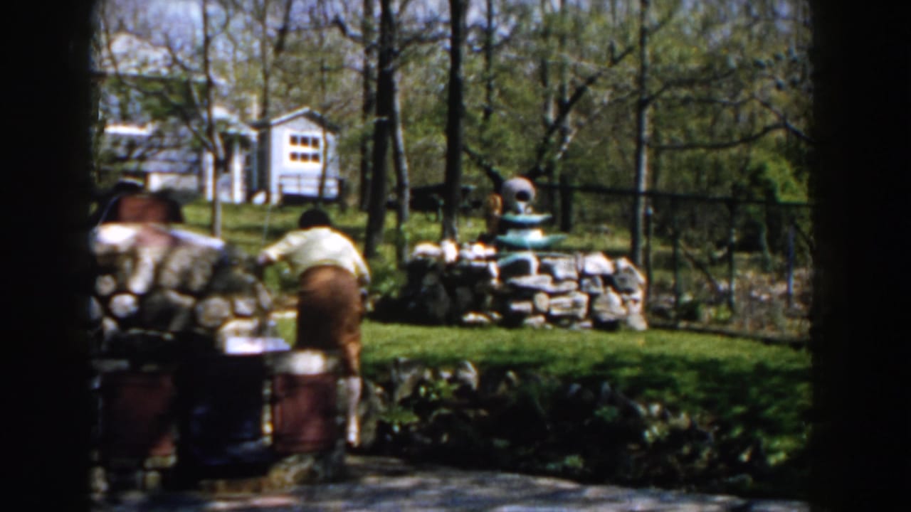 Woman near a stone garden fountain with trees and a small white structure behind her.