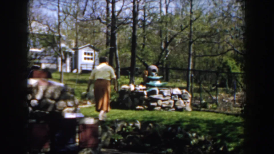 1959 Michigan 8mm frame showing the woman near the garden fountain, with lawn, trees, stonework, and a white structure in the background.