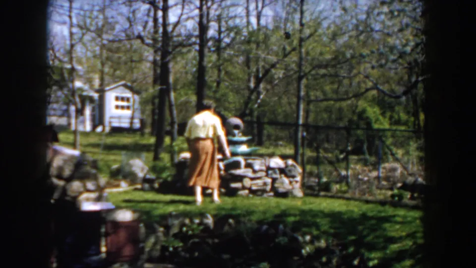 1959 Michigan home-movie frame showing the woman leaning near a stone garden fountain in a leafy backyard setting.