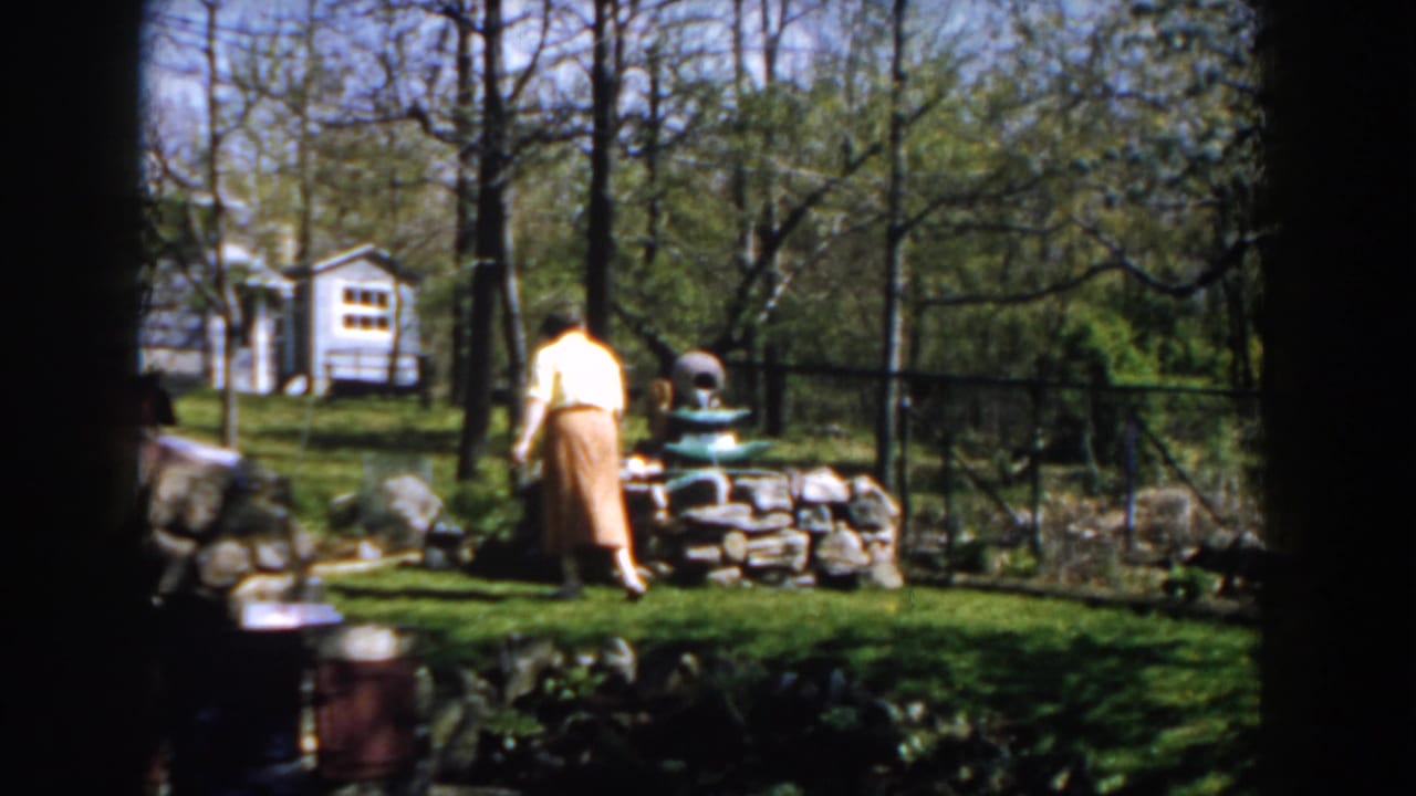 Woman in a yellow blouse and brown skirt stands near a stone garden wall in a sunny backyard.
