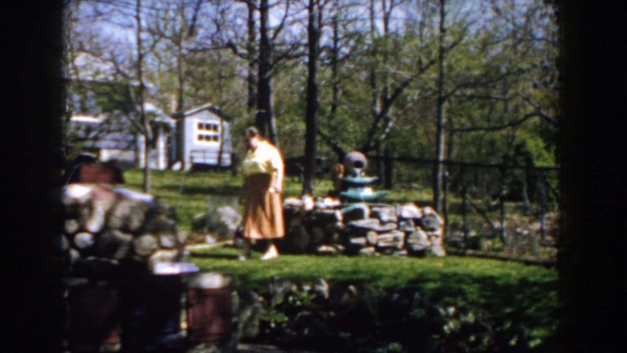 Woman walks across the grass past a stone wall and fountain in a leafy backyard.