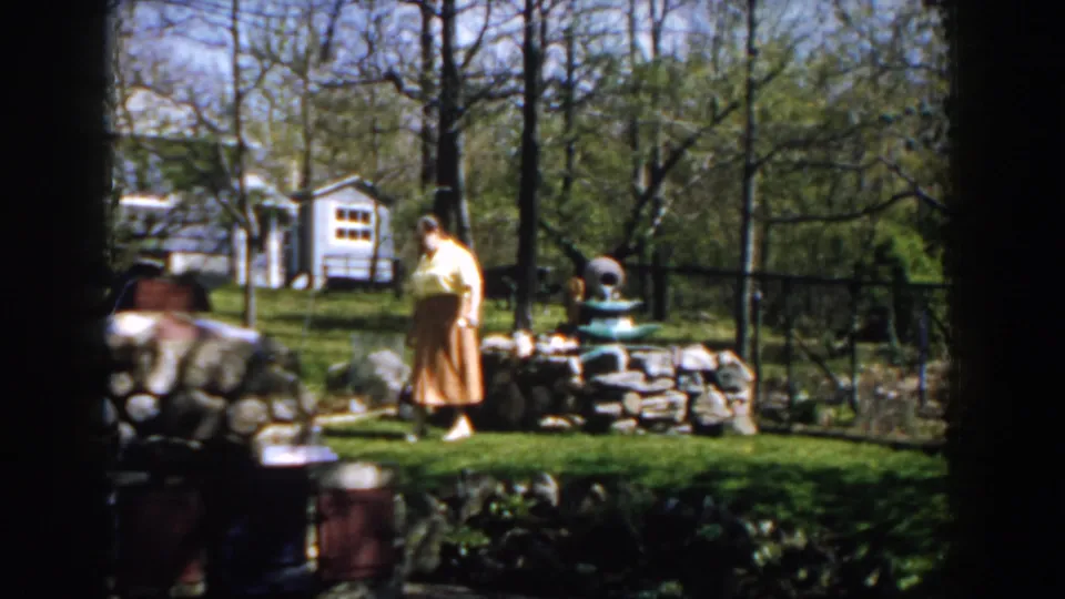 The woman walks through a sunny Michigan backyard, passing a low stone wall and garden fountain with trees and a white outbuilding beyond.