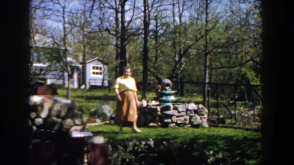 A woman in a yellow blouse and brown skirt faces the camera in a 1959 Michigan yard, with a stone border, fountain, trees, and white outbuilding visible.