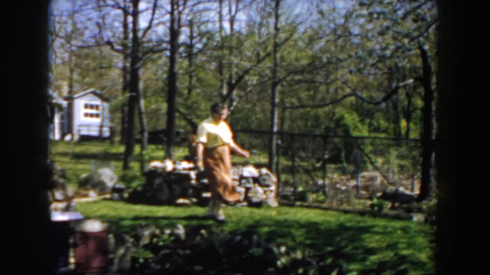 The woman continues across the lawn in 1959 Michigan footage, framed by a stone garden wall, fence line, trees, and a small white outbuilding.