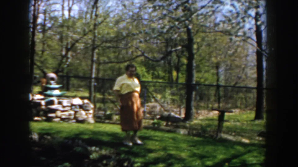 The woman walks slowly through a Michigan backyard, framed by lawn, trees, a low stone fountain, and a wire fence.