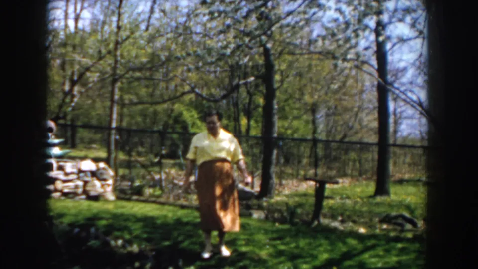 A woman in a yellow blouse and brown skirt walks closer through a green 1959 Michigan backyard, seen through dark framing.
