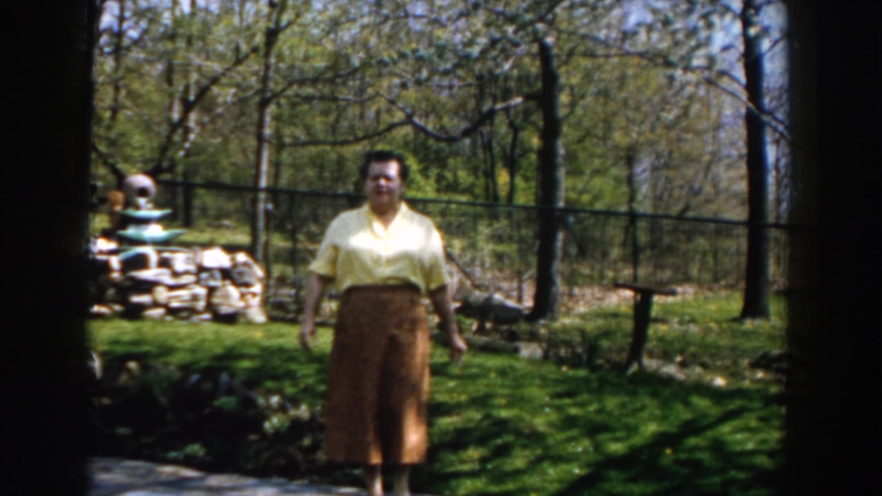 In 1959 Michigan home-movie footage, a woman in a yellow blouse and brown skirt faces the camera in a sunlit backyard with trees and a fence.