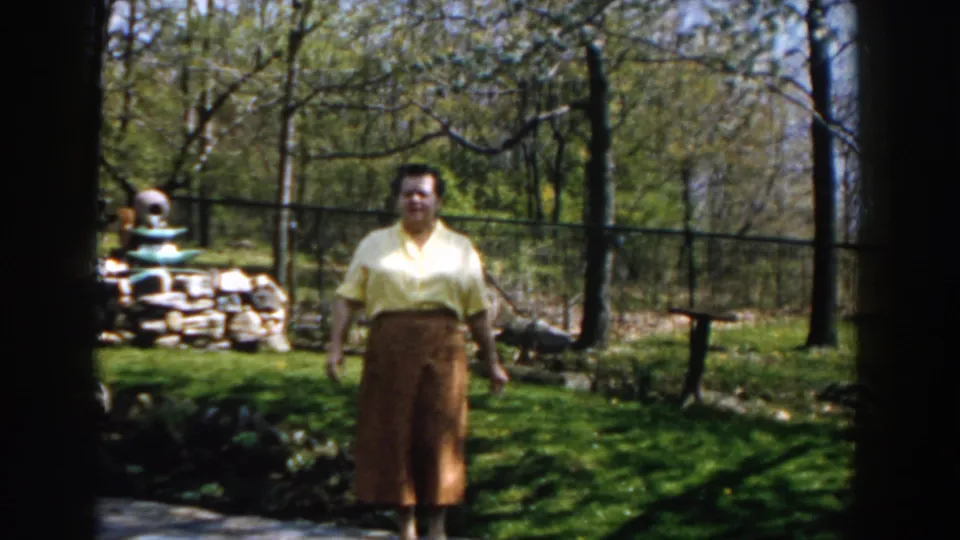 In 1959 Michigan home-movie footage, a woman in a yellow blouse and brown skirt faces the camera in a sunlit backyard with trees and a fence.