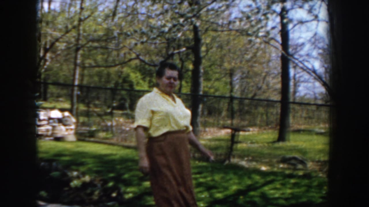 Woman walks across a grassy yard near trees and a fence, with sunlight falling across the lawn.
