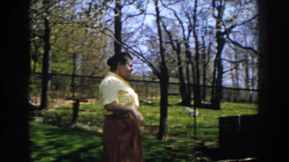 A profile view shows the woman gesturing in a 1959 Michigan backyard, framed by trees, grass, and a fence in soft 8mm color.
