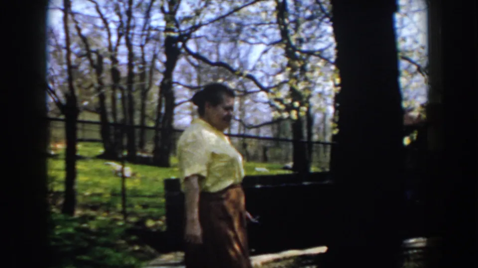 Near the end of the 1959 Michigan clip, the woman walks past a shadowed area of the backyard, with trees and fence lines behind her.