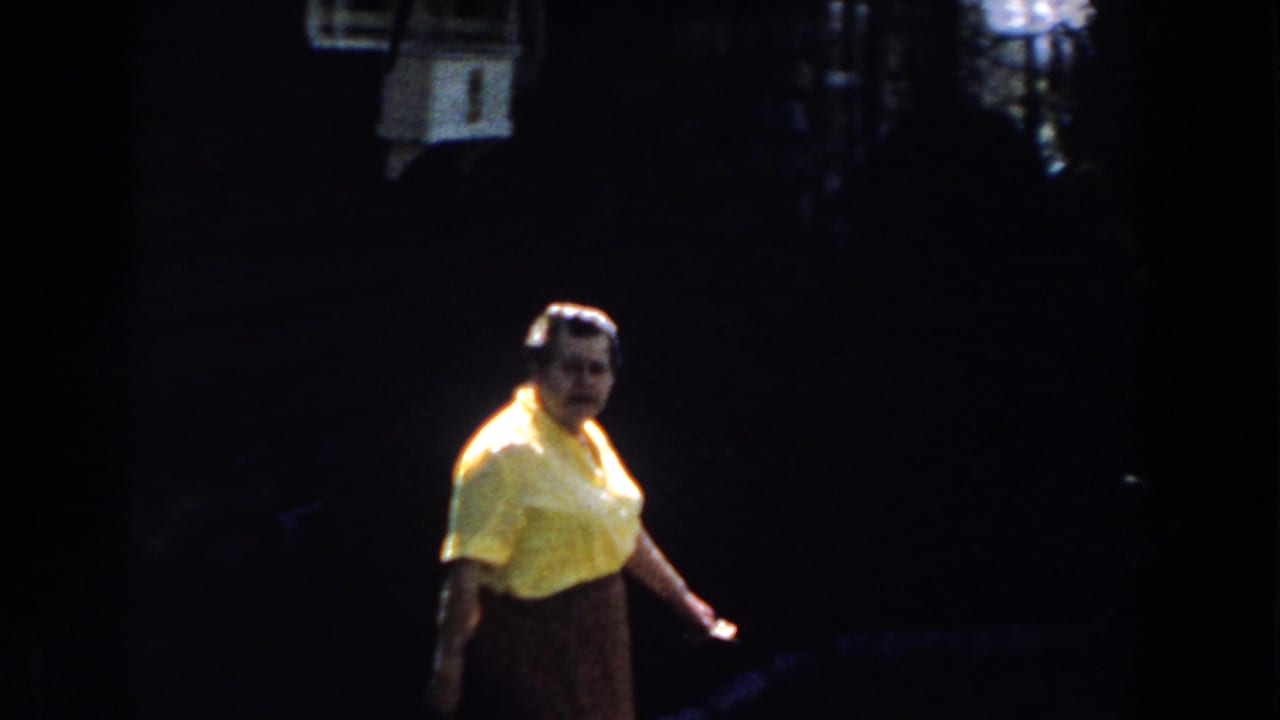 In 1959 Michigan home-movie footage, an adult woman in a yellow blouse and brown skirt walks near a shaded porch and lawn.