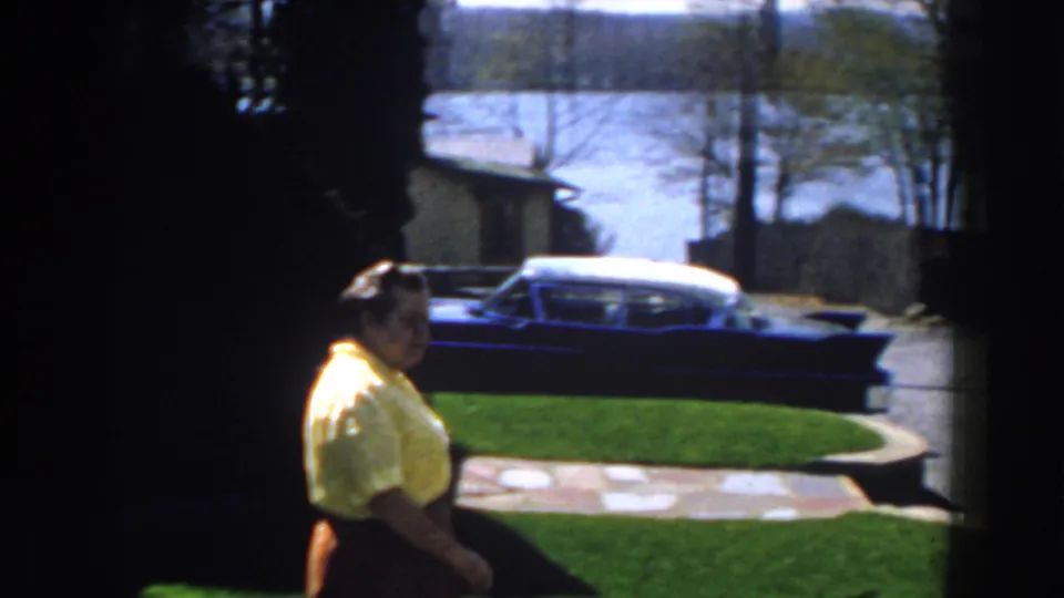 The woman moves through a suburban Michigan yard, with a mid-century sedan parked near the driveway in the background.