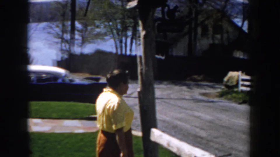 The woman continues away from the camera through the yard, with a driveway, trees, and houses visible in the bright background.