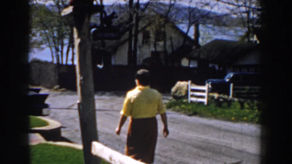 In 1959 Michigan home-movie footage, a woman walks away on a quiet street with houses, trees, a signpost, and lake water beyond.