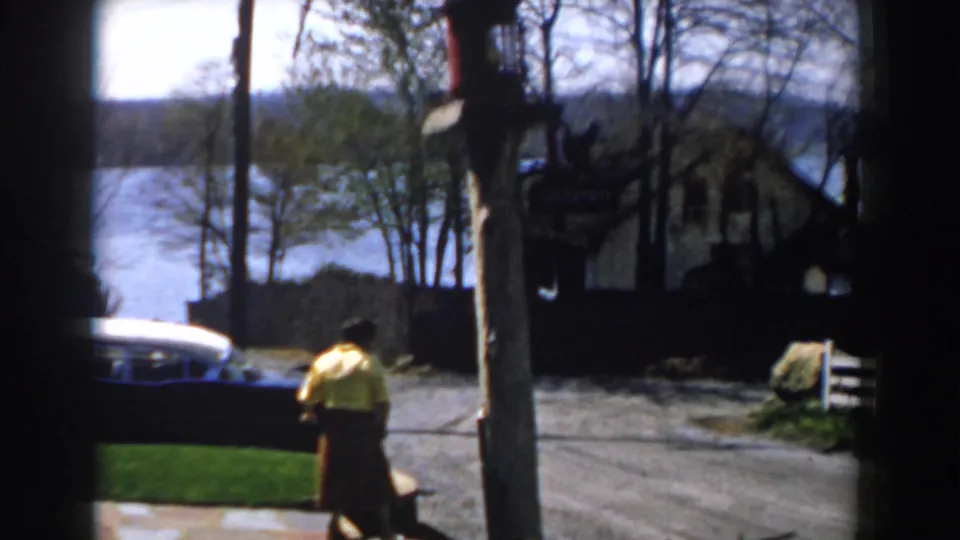 1959 Michigan 8mm frame showing a woman in a yellow shirt and red skirt near a lawn, parked car, signpost, trees, and water beyond the road.