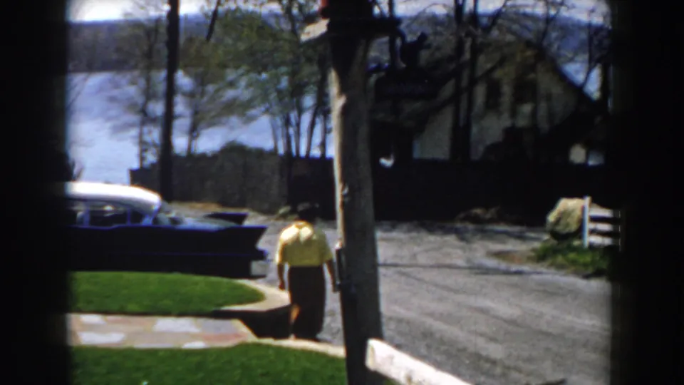 1959 Michigan home-movie frame with the woman moving away from the camera toward the road, seen near a parked blue and white car, signpost, trees, and lakeside background.