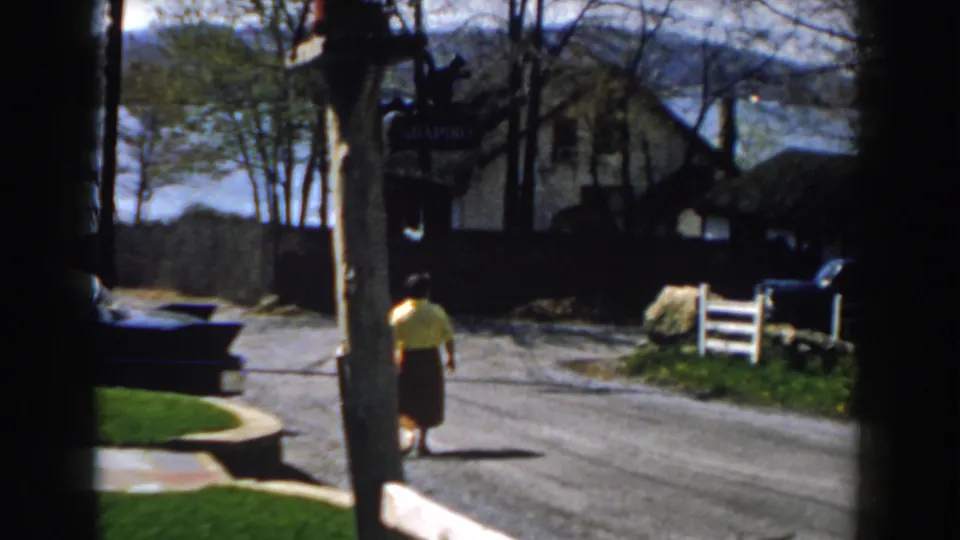 1959 Michigan home-movie frame showing the woman farther from the camera on the street, with a wooden sign, trees, parked vehicle, lawn, house, and water behind her.