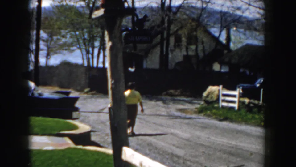 1959 Michigan 8mm frame showing an older woman walking on a sunlit residential street near a dark car, trees, a white fence, and water in the distance.